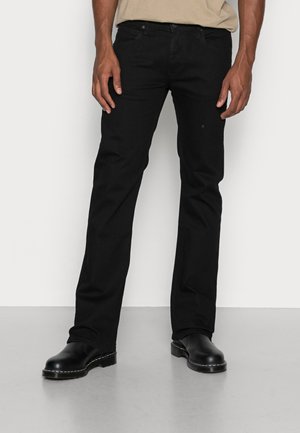 Man wearing black straight-leg jeans and black leather boots standing against a white background.