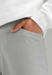 Pantaloni gri dintr-o țesătură elastică, cu un design elegant, având un buzunar pe lateral și cusături vizibile. Purtați cu un top alb cu mânecă lungă.