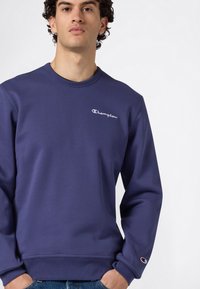 Joven con sudadera azul marino de Champion, con el logo en el pecho izquierdo y en la manga, con las manos en los bolsillos, contra un fondo liso.