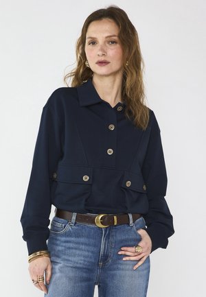 Femme aux cheveux châtain clair ondulés, portant une veste boutonnée bleu marine, un jean bleu, des bijoux en or et une ceinture marron avec une boucle en or.