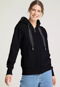 Schwarze Zip-Hoodie mit Kapuze, grauen Kordeln und Fronttaschen. Weicher Stoff, lässige Passform, kombiniert mit hellblauen Jeans.
