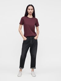 Femme aux longs cheveux noirs portant un t-shirt bordeaux, un jean noir à boutons et revers, et des baskets blanches, debout les mains dans les poches.