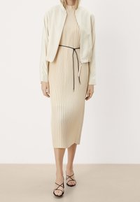 Abito midi plissettato beige con colletto alto, abbinato a una giacca bomber crema. Modella che indossa sandali neri con cinturini.
