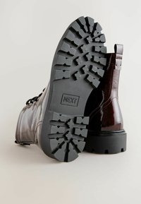 Botas de ante de cuero marrón brillante hasta el tobillo con una suela de goma negra gruesa que presenta un patrón de banda de rodadura rugoso y una lengüeta en la parte posterior.