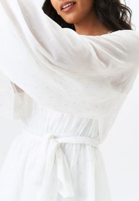 White sheer dress with textured pattern, billowy sleeves, and a cinched waist. Features a tie belt for adjustment. Lightweight fabric.