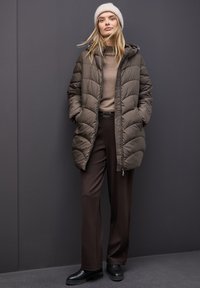 Abrigo acolchado marrón con bolsillos laterales y capucha, llevado sobre un suéter beige y pantalones marrones. El modelo lleva un gorro de punto de color claro.