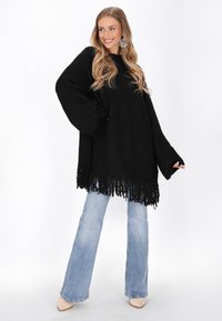 Schwarzer, übergroßer Pullover mit fransigem Saum, kombiniert mit hellblauen, weit geschnittenen Jeans und beigen Absätzen. Der Pullover hat einen strukturierten Stoff und weite Ärmel.