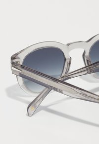 Transparente Sonnenbrille mit grauen Verlaufslinsen, kantigen Rahmen und strukturierten grauen Bügeln mit feinen Linien und einem goldenen Logo-Akzent.