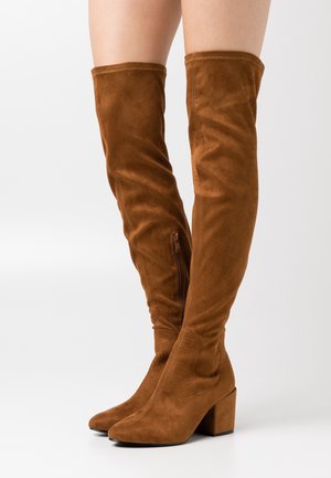 Bottes en daim marron au-dessus du genou avec talons carrés et fermetures éclair latérales, portées sur des jambes nues sur un fond blanc uni.