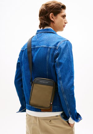 Mand iført blå denimjakke og beige bukser bærer en olivengrøn Tommy Hilfiger skuldertaske med sort rem.