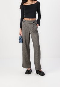 Schwarzes, langärmeliges, cropped Oberteil und weit geschnittene, gemusterte Hosen in Braun und Beige. Die Hosen haben seitliche Taschen und Knopfdetails. Schwarze, klobige Schuhe.