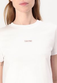 Tricou alb din bumbac cu guler rotund, având un mic patch cu logo-ul roșu și alb Calvin Klein pe față.