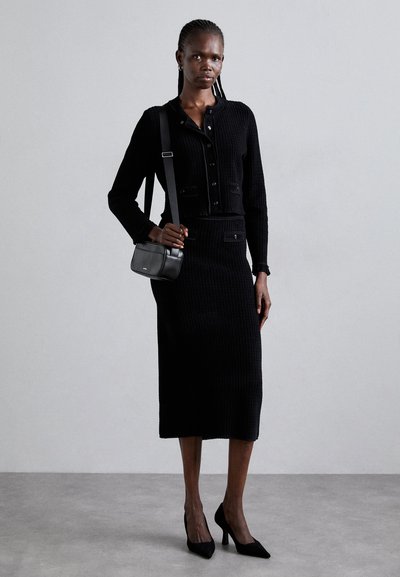 Mustaan neulottu kahden osan asu, joka koostuu lyhyestä napitettavasta yläosasta ja tiukasta midi-hameesta. Yhdistettynä pieneen mustaan olkalaukkuun ja korkokenkiin.