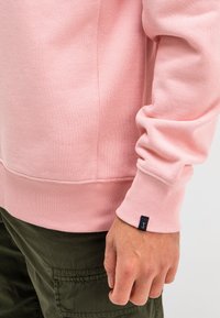 Roze sweatshirt met ribgebreide manchetten en zoom, met contrasterende stiksels en een klein zwart logo-label op de manchette. Draag het met groene broek.