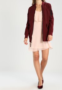 Burgundská bomber bunda se zipem a boční kapsou, nošená přes světle růžové plisé šaty s volánovým lemem. Tmavě červené podpatky.