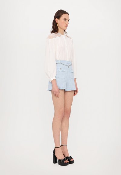 Blusa blanca con detalles de encaje en los hombros, combinada con pantalones cortos de tweed azul claro que tienen bolsillos y una cadena decorativa. Sandalias de plataforma negras.