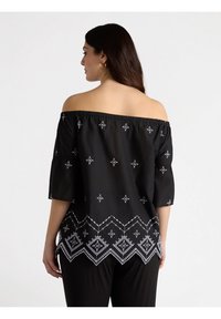 Top negro fuera del hombro con intrincados bordados blancos y mangas cortas. Presenta un dobladillo con patrón en zigzag y diseños geométricos.