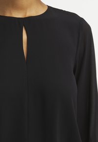 Nainen, joka on pukeutunut mustaan pitkään hihaan olevaan puseroon, jossa on pieni pystysuora avaus kaula-aukossa.