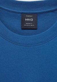 Modrá bavlnená tričko má okrúhly výstrih, rebrovaný golier a štítok s nápisom "ESENCIÁLY MNG REGULÁRNA VEĽKOSŤ EUR-USA M / MEX M."