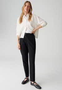 Femme debout portant une chemise blanche texturée à boutons, un pantalon noir sur mesure et des ballerines Mary Jane noires, sur un fond gris uni.