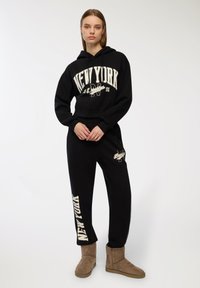 Czarny sweatshirt z kapturem z grafiką "NEW YORK" w odcieniu kremowym, w zestawie z czarnymi spodniami dresowymi i brązowymi zamszowymi botkami do kostki. Zauważalna gładka фактурa materiału.