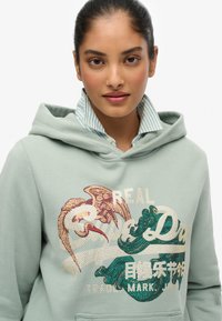 Giovane donna che indossa una felpa verde chiaro con un grafico di un uccello e un'onda, e una camicia a righe a strati sotto, sfondo neutro.