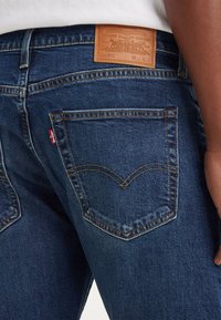 Dunkelblaue Jeans im klassischen Schnitt. Zu den Merkmalen gehören gestickte Gesäßtaschen, ein Leder-Logoaufnäher und sichtbare Nähte.