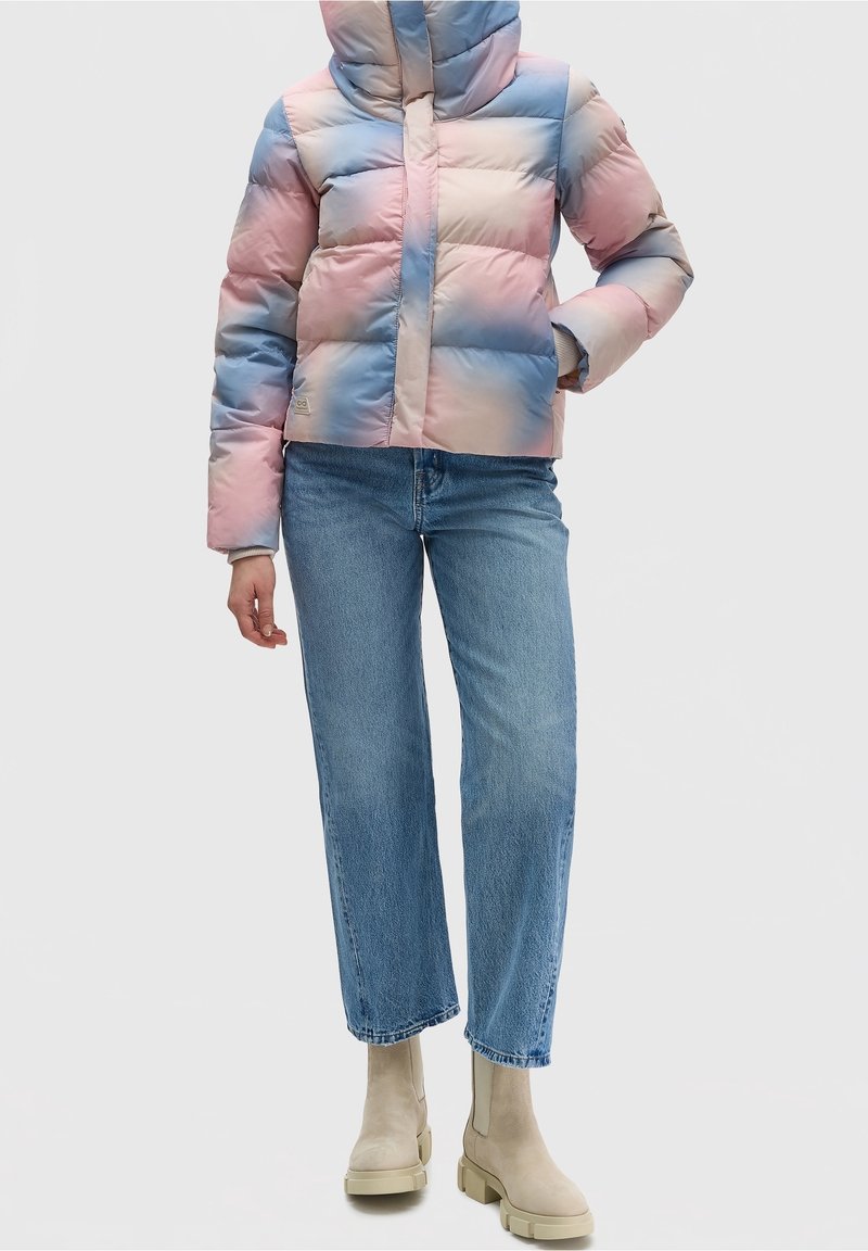 Femme portant une veste matelassée rose pastel et bleue, un jean droit bleu clair, et des bottines à semelle épaisse beige, se tenant devant un fond blanc.