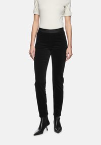 Schwarze Samthose mit einem taillierten Design und einem strukturierten Taillenbund. Getragen mit einem cremfarbenen Oberteil und schwarzen High-Heels.