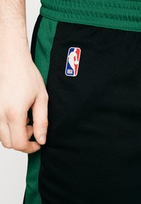 Basketballshorts lavet af åndbart mesh-stof, med sort som primærfarve og grønne accenter, samt et NBA-logo patch på siden.