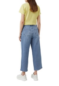 Světle žlutý top s krátkým rukávem v kombinaci s vysokým pasem širokými modrými džínami a bílými teniskami, s zadními kapsami a volným střihem.