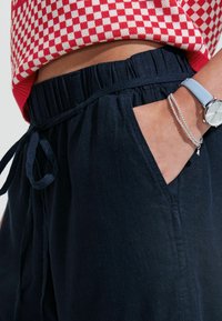 Tamsiai mėlynos lininės kelnės su plačia elastine juosta, šnervimo raiščiu ir šoninėmis kišenėmis; derinamos su raudonai languotu megztiniu.