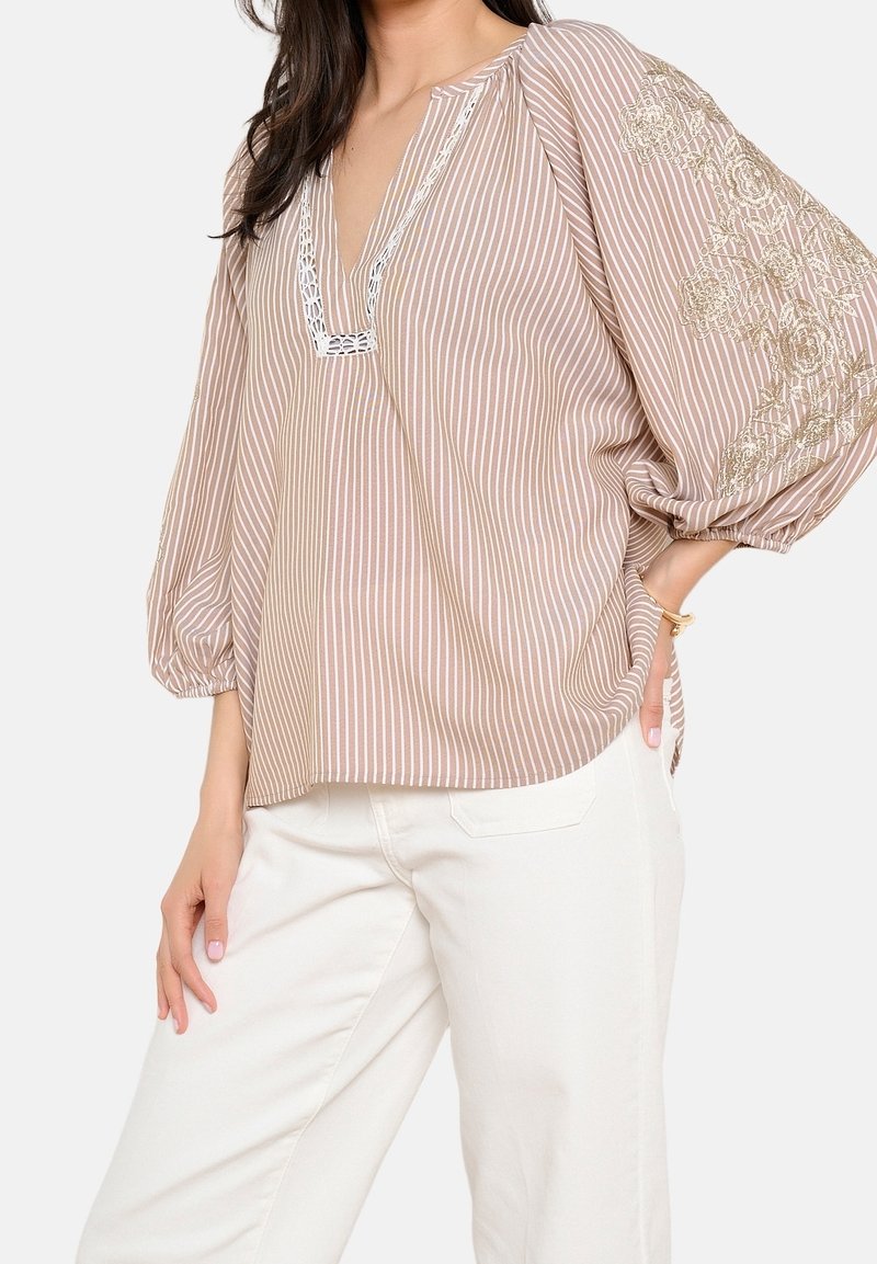 Femme portant un chemisier beige et blanc à rayures avec des motifs floraux brodés sur des manches bouffantes et un pantalon blanc, main posée sur la hanche.