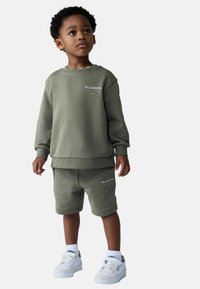 Oliivinvihreä puuvillainen collegepaita ja shortsit, joissa on logokoristeita. Tennarit ovat valkoiset harmailla yksityiskohdilla. Yksinkertainen, rento muotoilu.