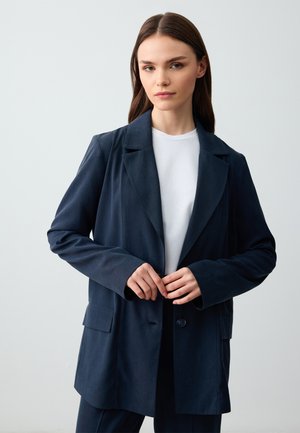 Navyblauwe blazer met een gegroefd kraag, enkele knoopsluiting, voorkruiszakken en lange mouwen, gedragen over een wit shirt. Gladde stofstructuur.