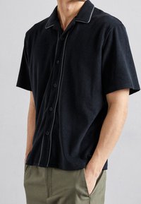 Lyhythihainen musta napillinen paita pehmeästä froteekudoksesta, jossa on valkoinen verkkokudontaviimeistely kauluksessa. Ominaisuuksiin kuuluu rento istuvuus ja hienovarainen logotieto.