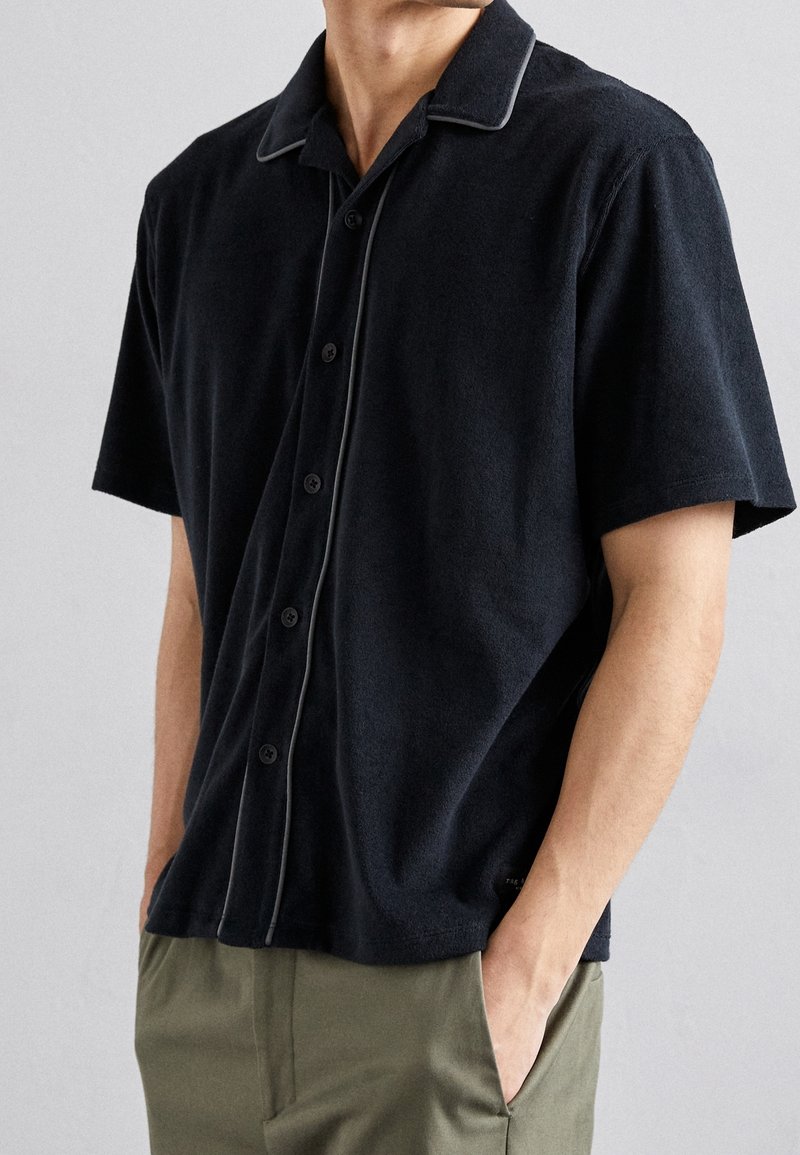 Lyhythihainen musta napillinen paita pehmeästä froteekudoksesta, jossa on valkoinen verkkokudontaviimeistely kauluksessa. Ominaisuuksiin kuuluu rento istuvuus ja hienovarainen logotieto.