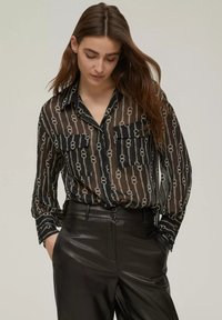 Blusa nera trasparente con motivo a catena argento, maniche lunghe, collo con bottoni, due tasche frontali, indossata con pantaloni in pelle neri a vita alta.