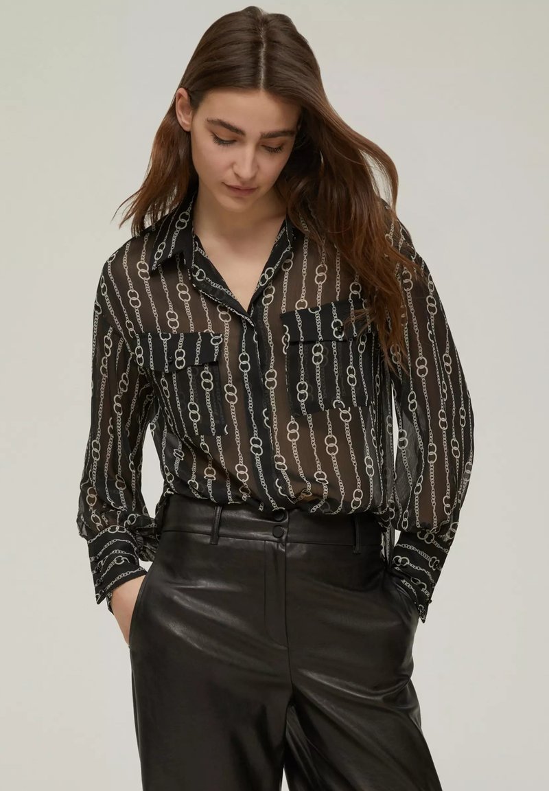 Blusa nera trasparente con motivo a catena argento, maniche lunghe, collo con bottoni, due tasche frontali, indossata con pantaloni in pelle neri a vita alta.