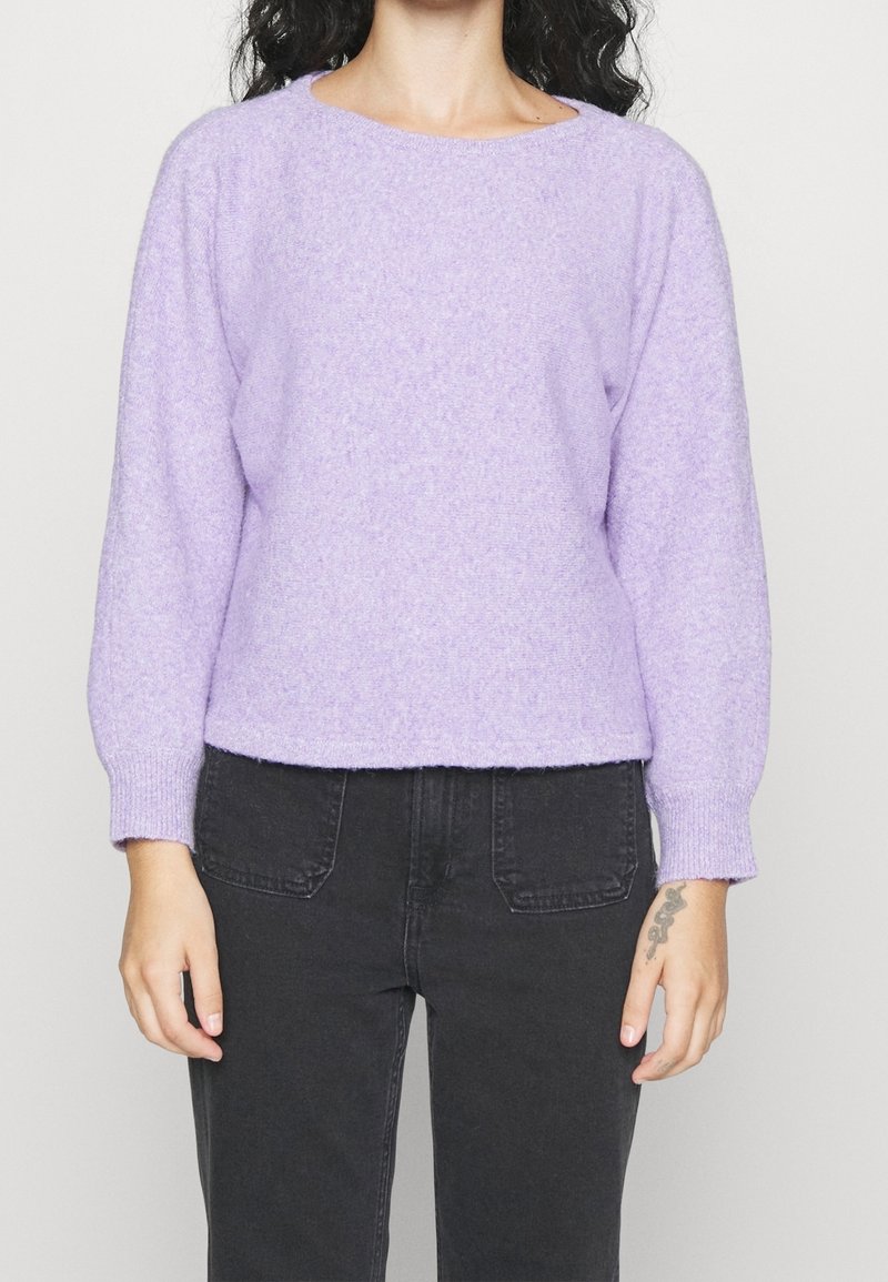 Femme portant un pull violet lavande à manches longues et duveteux ainsi qu’un jean noir, avec un petit tatouage visible sur sa main droite.