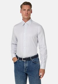 Weißes Hemd mit blauen vertikalen Streifen; lange Ärmel mit Knopfmanschetten. Kombiniert mit blauen Jeans und einem geflochtenen schwarzen Gürtel.