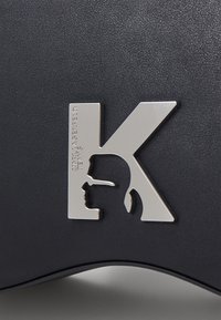 Materiale nero testurizzato con un elegante logo in metallo argento a forma di "K" stilizzata con un dettaglio della silhouette profilo.