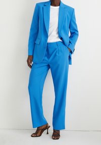 Costume bleu vif tailleur avec un blazer structuré et un pantalon ample, associé à un t-shirt blanc et des talons noirs. Texture de tissu lisse.