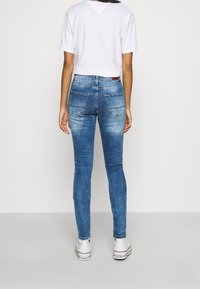 Jeans skinny de mezclilla azul con efecto desgastado, cinco bolsillos y un parche de cuero en la cinturilla trasera. Combinados con zapatillas blancas.