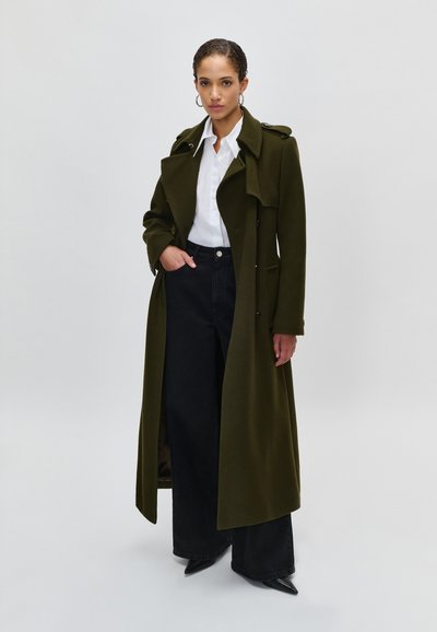 Mulher vestindo um casaco longo verde-oliva, camisa branca com botões, calças largas pretas e sapatos pretos de bico contra um fundo simples.