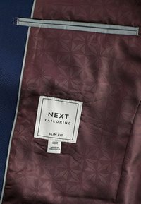 Slim-Fit-Jacke mit Innenfutter aus dunkelblauem strukturierten Stoff und bordeauxroter Fütterung, die ein geometrisches Muster aufweist, sowie einem weißen Etikett.