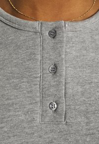 Primo piano di una persona che indossa una camicia grigia con tre bottoni visibili e una sottile collana d'oro sul colletto.