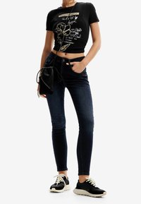 Top corto negro con diseño gráfico, jeans ajustados azul oscuro y zapatillas negras. Pequeño bolso negro sostenido en una mano.