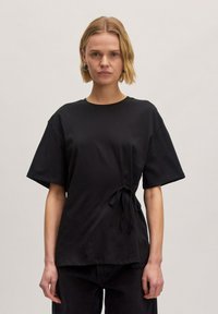 Zwarte kortemouwenshirt van zachte stof met een losse fit en een zijstrikdetail. Ronde halslijn en minimalistisch ontwerp.