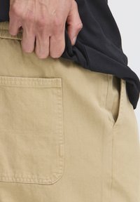 Shorts en coton couleur fauve avec une poche latérale et un tissu texturé. Chemise noire partiellement rentrée. Main saisissant le tissu de la chemise.